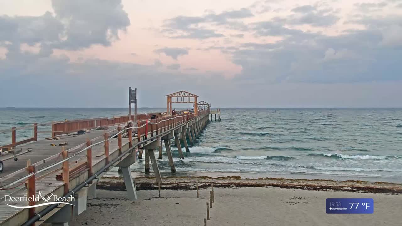 A partially constructed wooden pier extends over a gently wavy ocean under a cloudy, pale sky.
