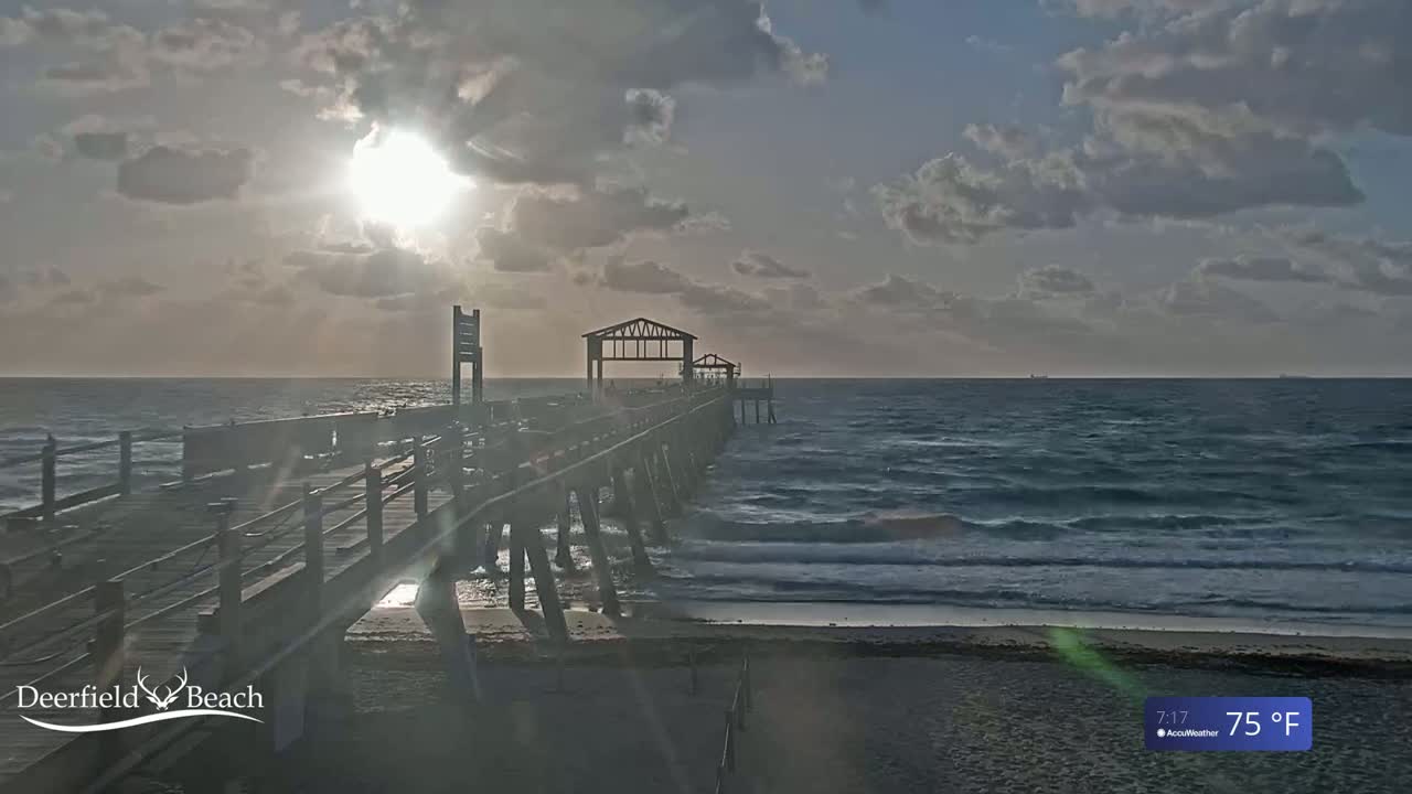 A long wooden pier extends into a partly cloudy ocean under a bright sun, with gentle waves lapping the sandy shore.