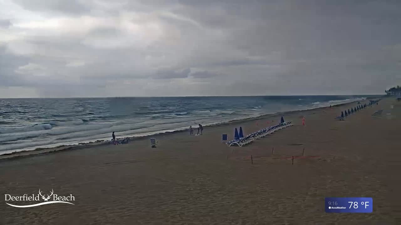 A mostly cloudy day shows a long, sandy beach with a few people and rows of lounge chairs facing a somewhat choppy ocean.