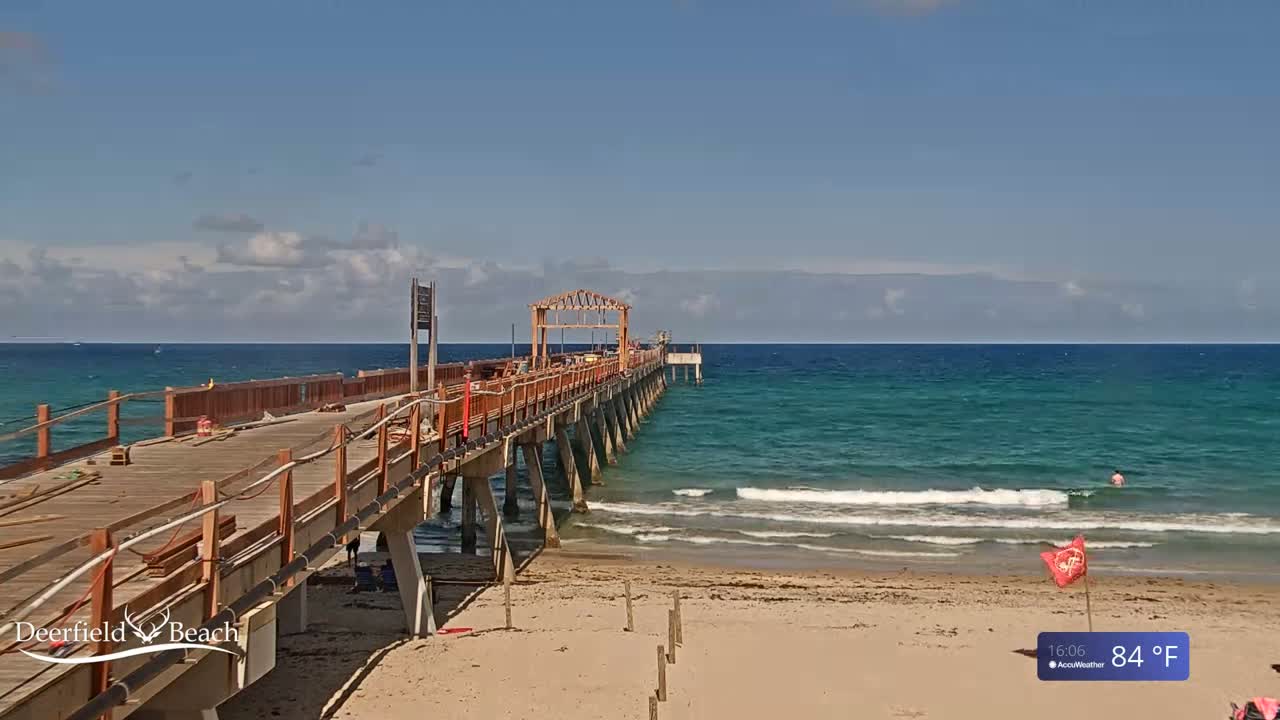 A long wooden pier extends over a teal ocean under a partly cloudy sky, with a sandy beach in the foreground on a 84°F day.