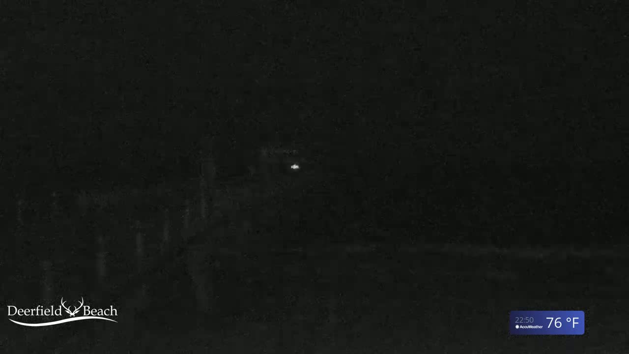 A dimly lit nighttime scene shows a dark expanse of land, possibly a beach, with a single, faint light visible in the distance under a dark sky.