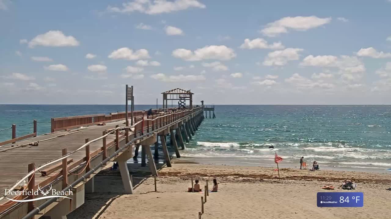 A partly cloudy day shows a wooden pier extending over a blue ocean with light waves and a sandy beach with a few people.