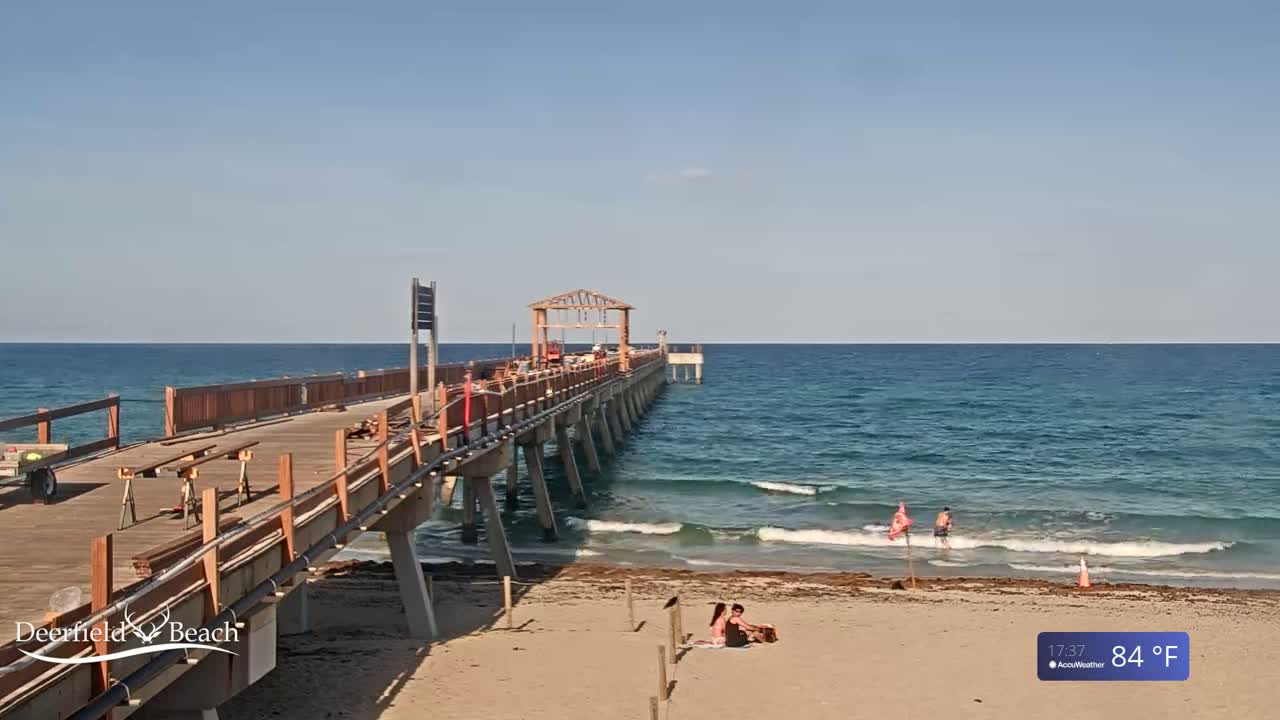 A long wooden pier extends over a calm, blue ocean on a sunny 84°F day, with a few people visible on the beach and in the water.