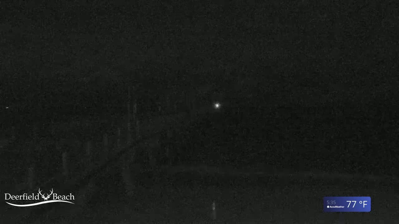 A nighttime, low-light image shows a dark, possibly wooded area under a clear sky, with a single bright light visible in the distance.