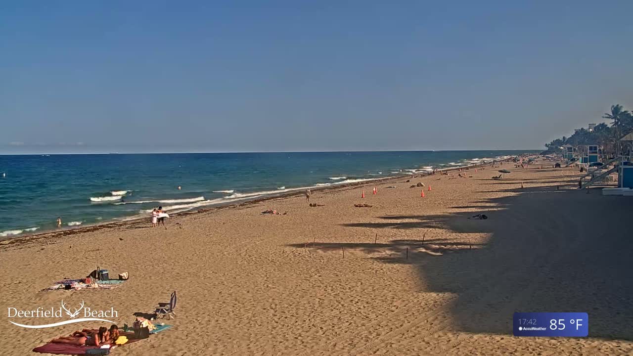 A sandy beach with a few people sunbathing and swimming in a calm ocean under a clear blue sky on a sunny 85°F day.