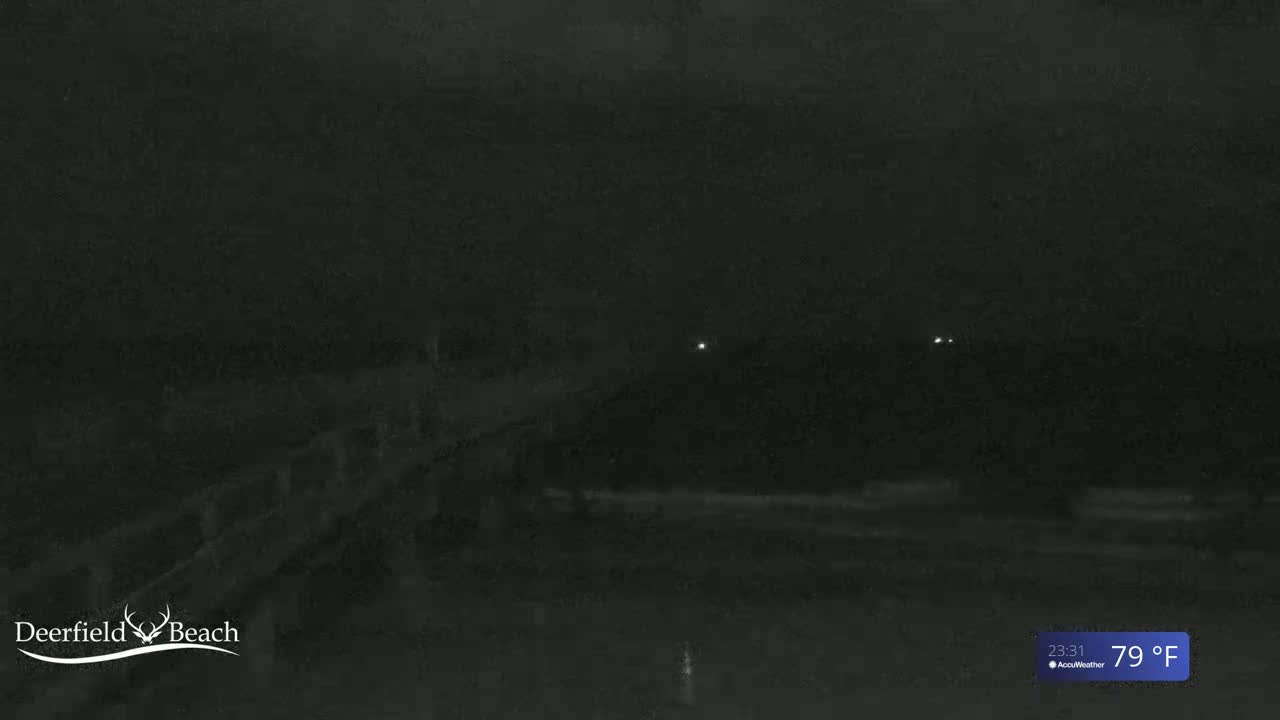 A nighttime, low-light view of a dark beach at 79°F, showing a dimly lit structure in the foreground and faint lights in the distance.