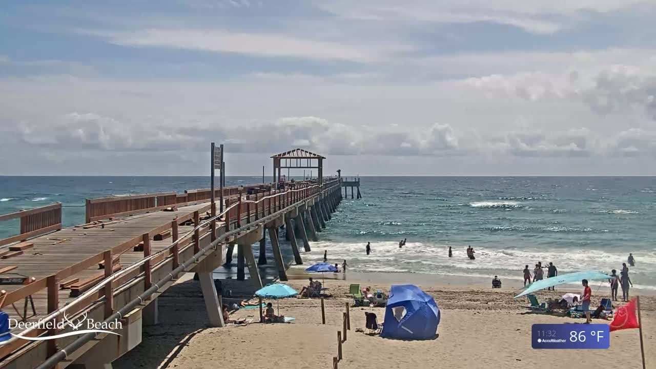 A partly cloudy day shows a long wooden pier extending over a gently wavy ocean, with people swimming and relaxing on the beach below.