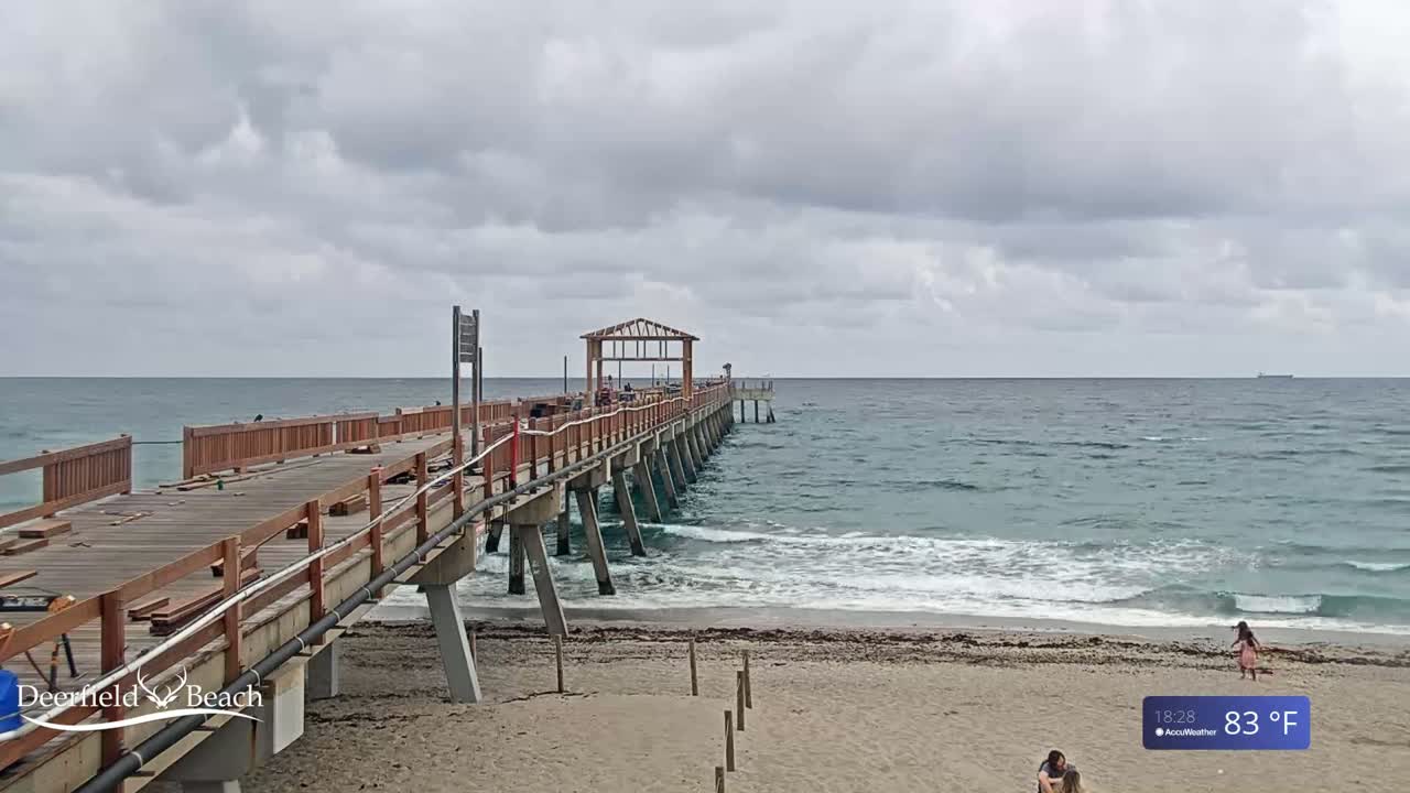 A wooden pier extends from a sandy beach into a calm ocean under a cloudy sky.