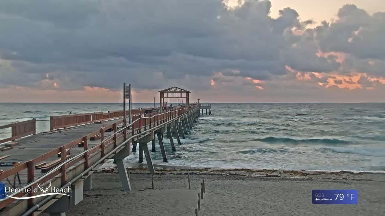 A long wooden pier extends over a wavy ocean under a partly cloudy sky at sunrise.