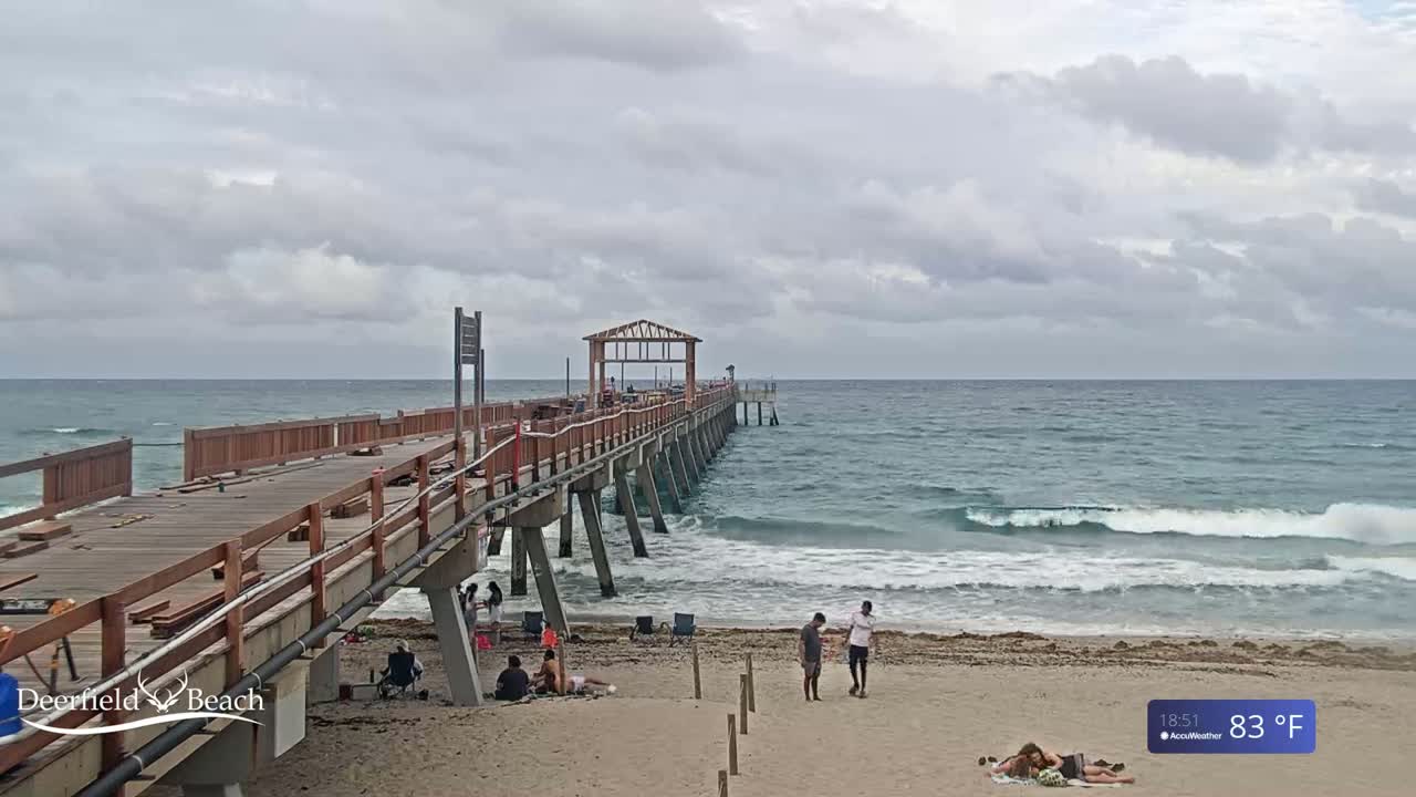 A partly cloudy day shows a long wooden pier extending over a beach with small waves, and several people are visible on the beach and the pier.