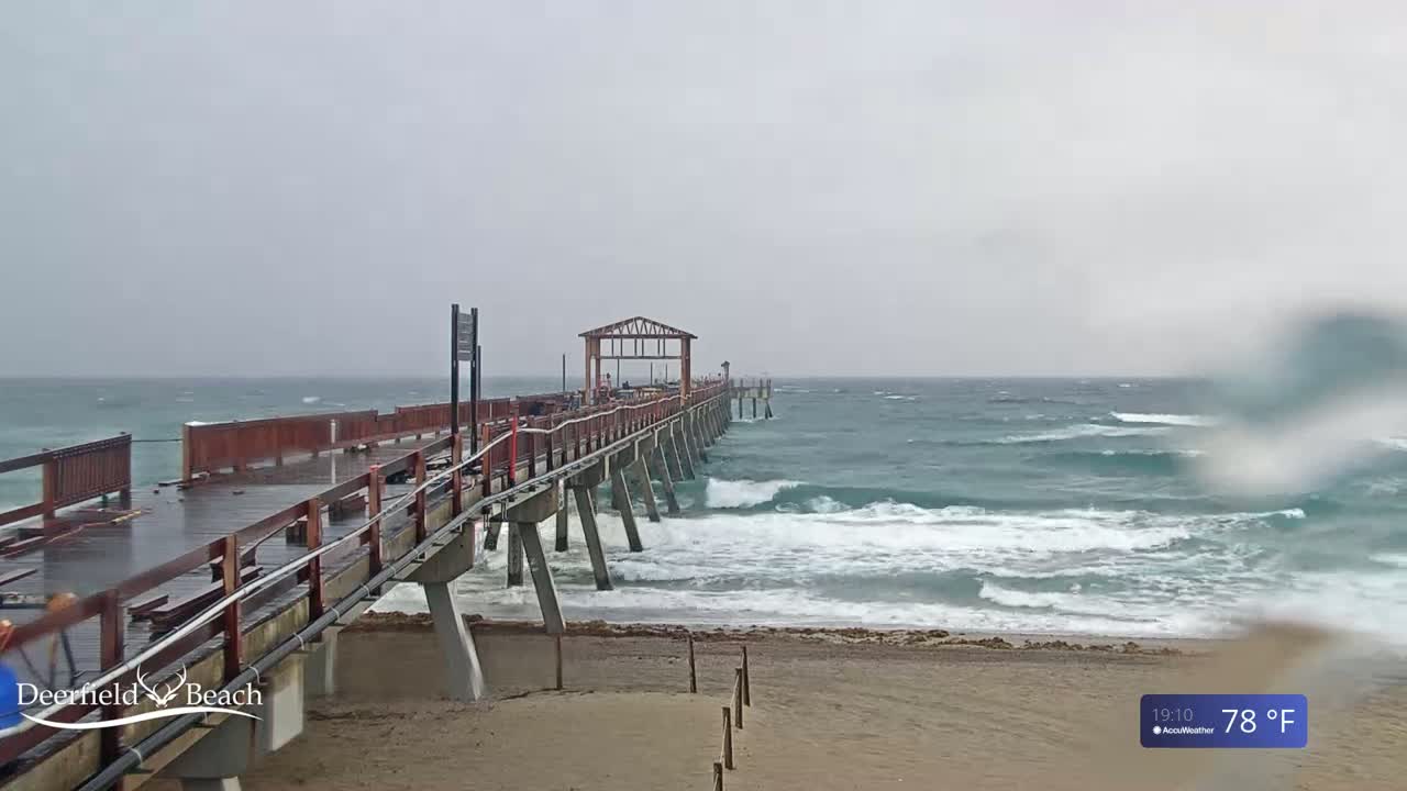 A long wooden pier extends over a wavy ocean under an overcast sky.