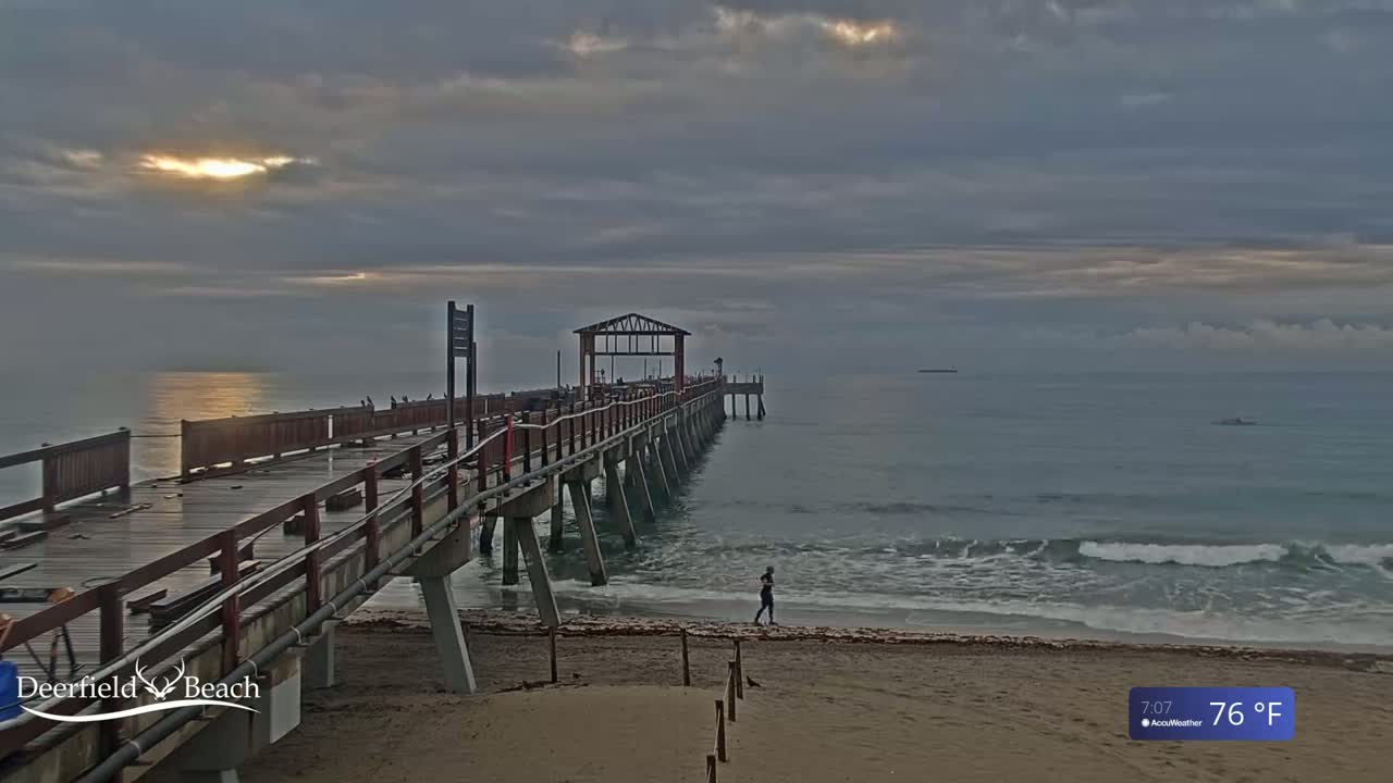 A long wooden pier extends into a calm ocean under a partly cloudy sky at sunrise.
