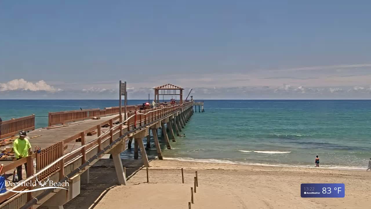 A partly cloudy, sunny day shows a long wooden pier under construction extending from a sandy beach into a calm, teal ocean.