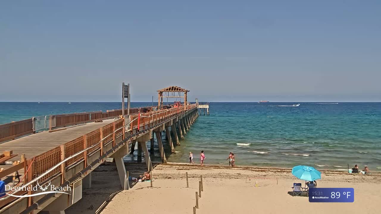 A long wooden pier extends over a calm, turquoise ocean on a sunny day, with a sandy beach and several people enjoying the warm weather.
