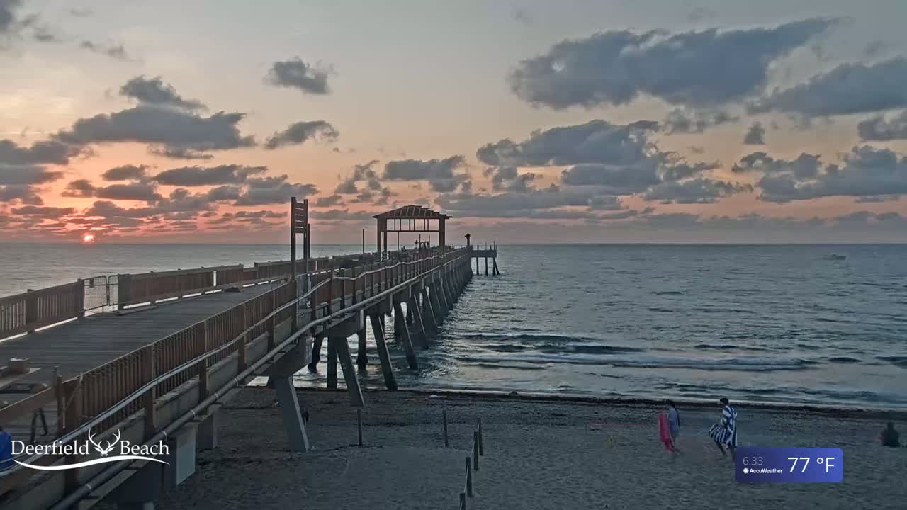 A long wooden pier extends over a calm ocean during a sunrise with partly cloudy skies, and a few people are visible on the beach.