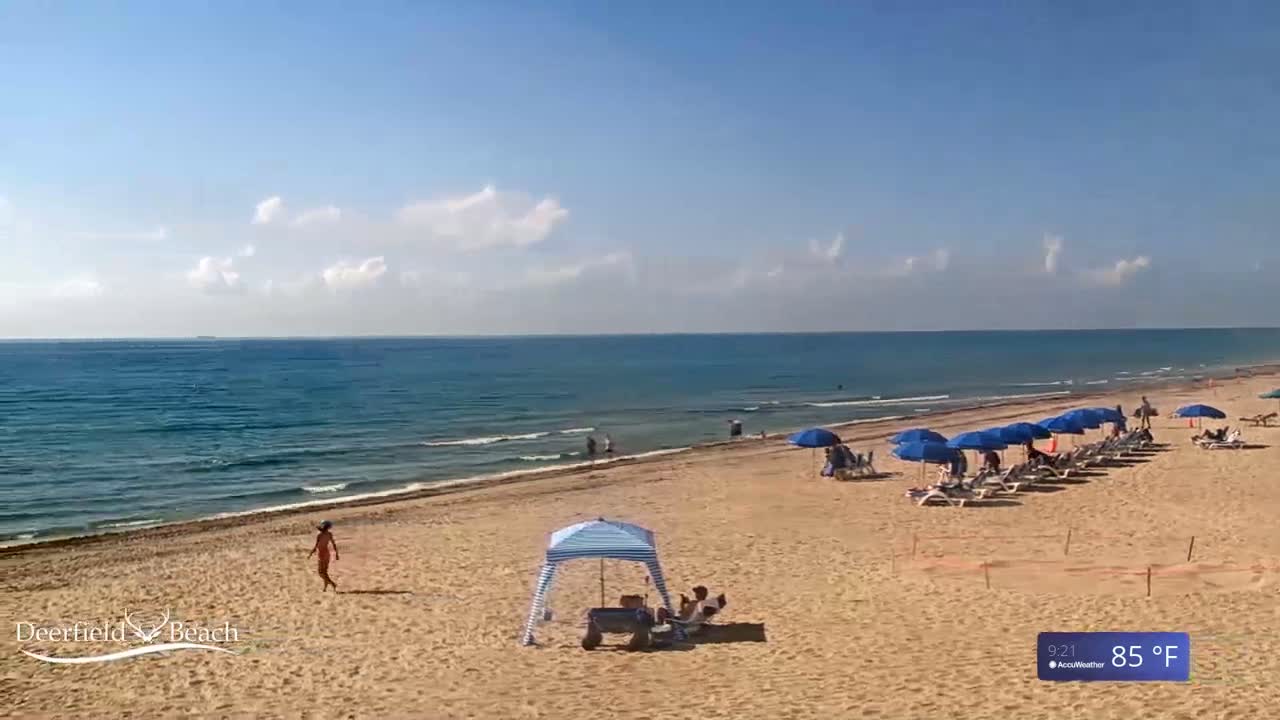 A sandy beach with several blue umbrellas and beach chairs under a mostly sunny sky, with calm ocean waves in the background.