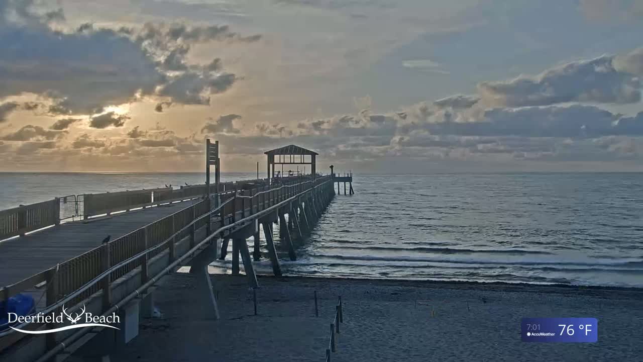 A long pier extends into a calm ocean under a partly cloudy sky at sunrise.