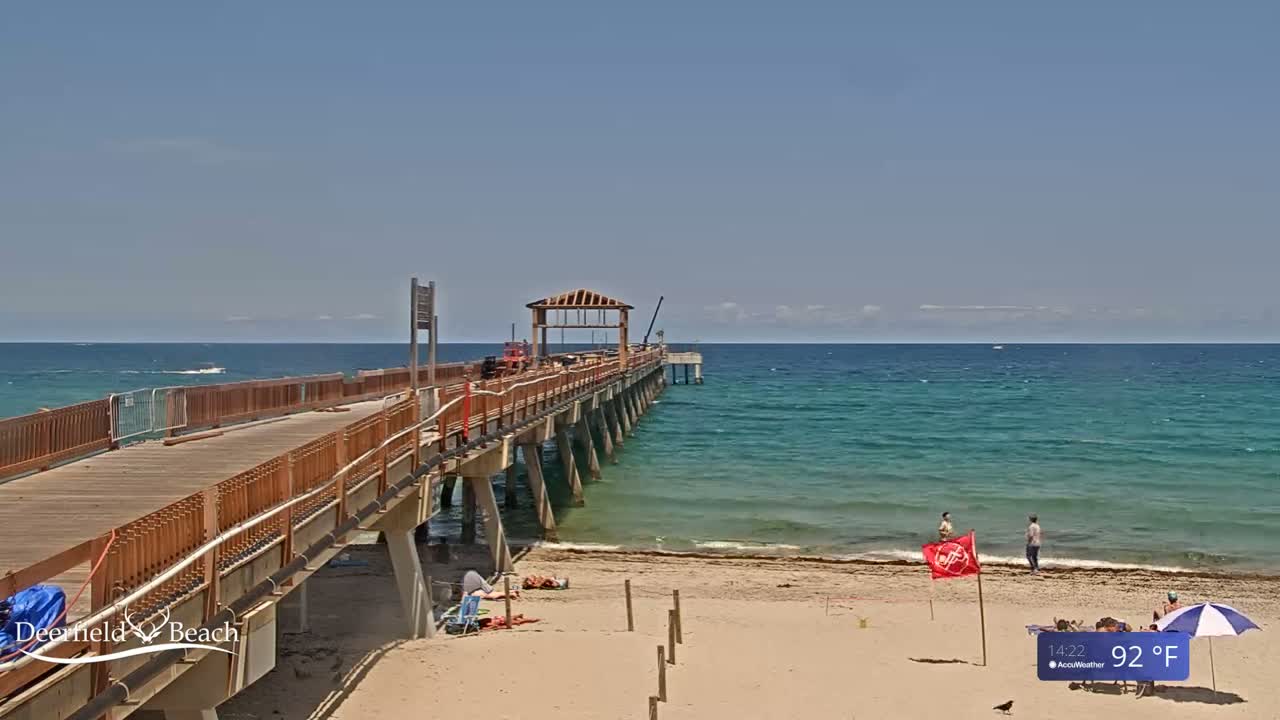 A long wooden pier extends over a calm, teal ocean on a sunny day with a temperature of 92°F.