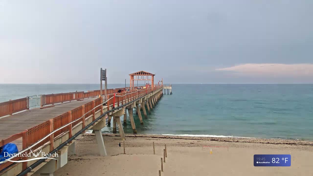 A long wooden pier extends over a calm, teal ocean toward a mostly cloudy sky, with a sandy beach visible below.