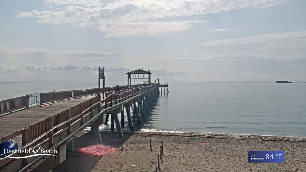 A long pier extends over a calm, light-blue ocean under a mostly cloudy sky, with a sandy beach visible in the foreground.
