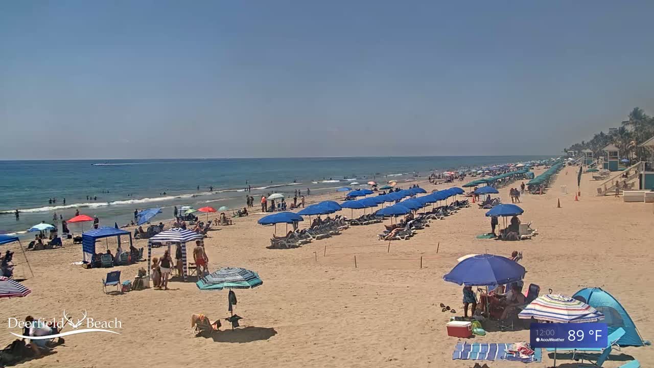 A sunny beach scene shows many people relaxing under umbrellas and beach tents on a sandy beach with calm ocean waves under a clear blue sky.