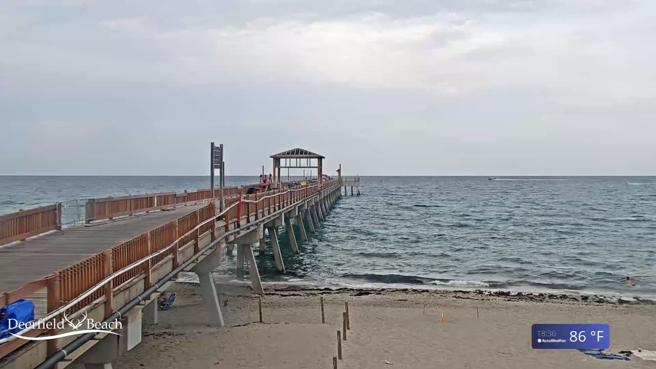 A long wooden pier extends over a calm ocean under an overcast sky, with a sandy beach visible in the foreground.