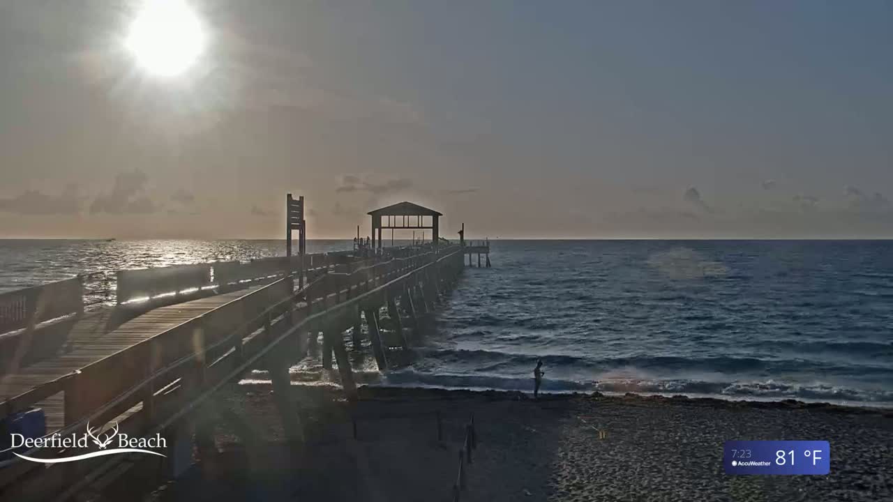 A long pier extends into a calm ocean under a bright, slightly hazy sun, with a sandy beach visible in the foreground and the temperature at 81°F.