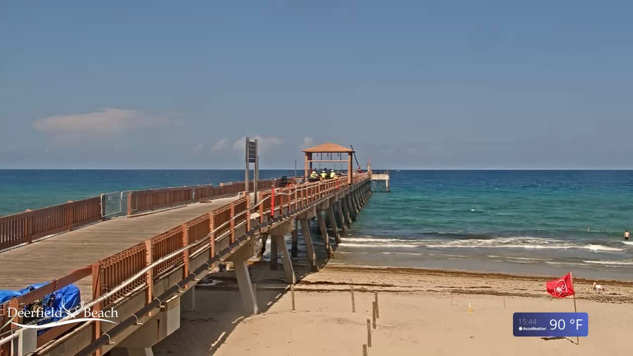 A long wooden pier extends from a sandy beach into a calm, teal ocean under a mostly clear blue sky.