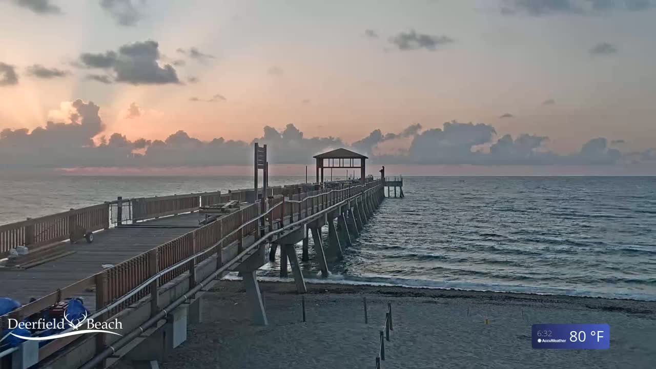 A long pier extends into a calm ocean under a partly cloudy, pinkish sunrise sky.
