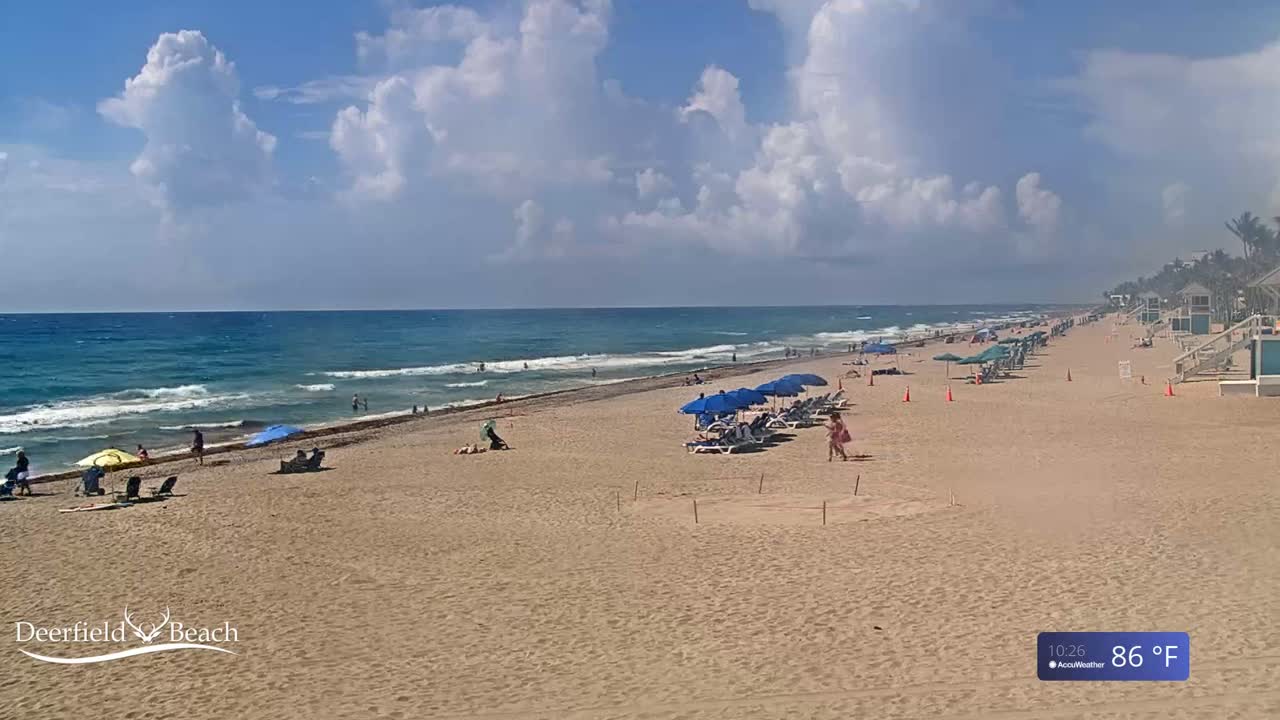 A sandy beach with blue umbrellas and lounge chairs stretches along a blue ocean under a partly cloudy sky.