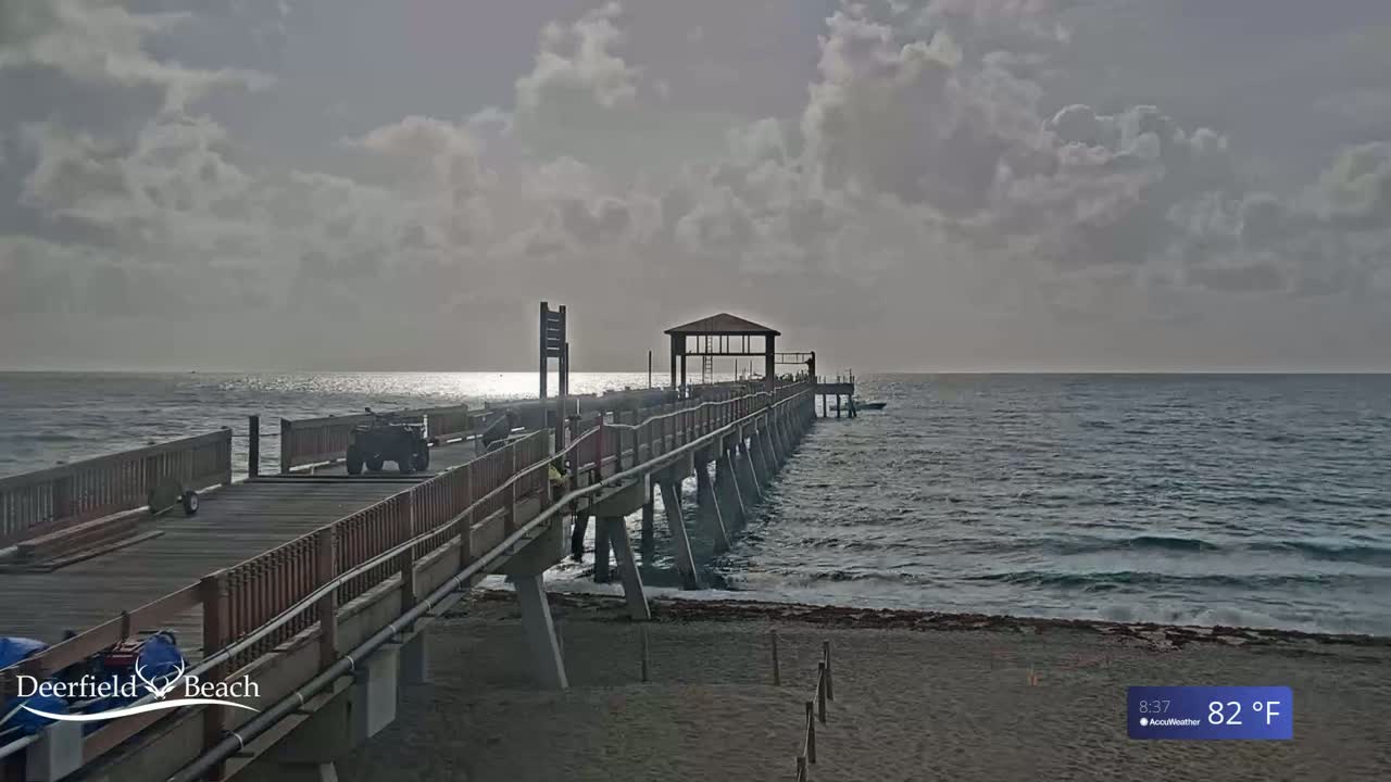 A long pier extends over a calm ocean under a cloudy sky.