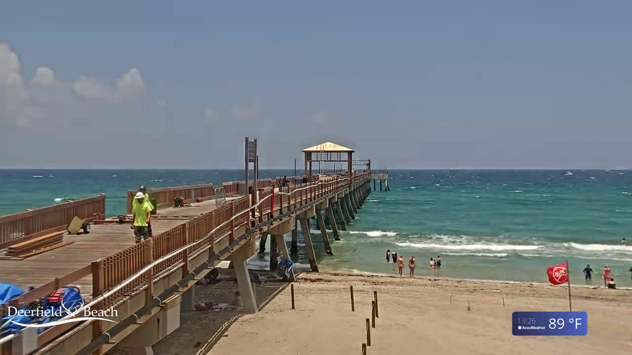 A long wooden pier extends over a teal ocean on a partly cloudy, 89°F day, with several people swimming in the water and construction workers on the pier.