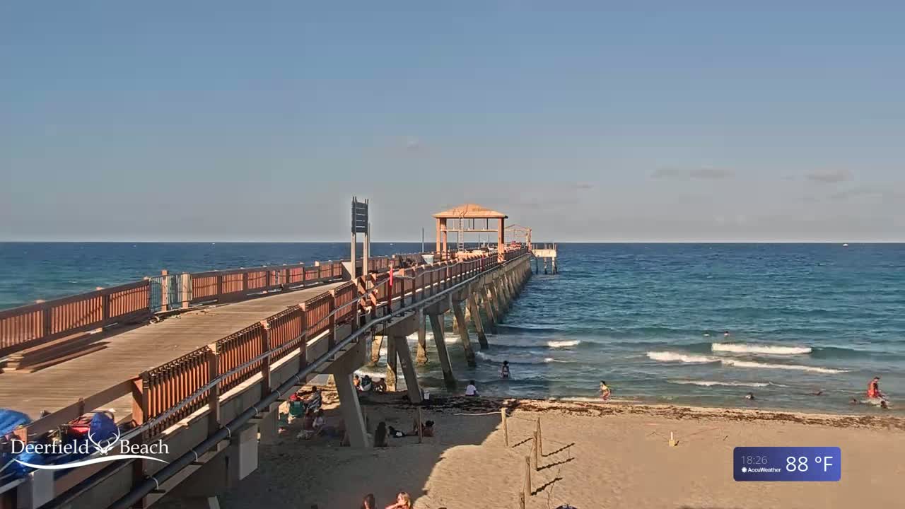 A long pier extends over a teal ocean with gentle waves under a partly cloudy sky, with people visible on the beach and the pier.