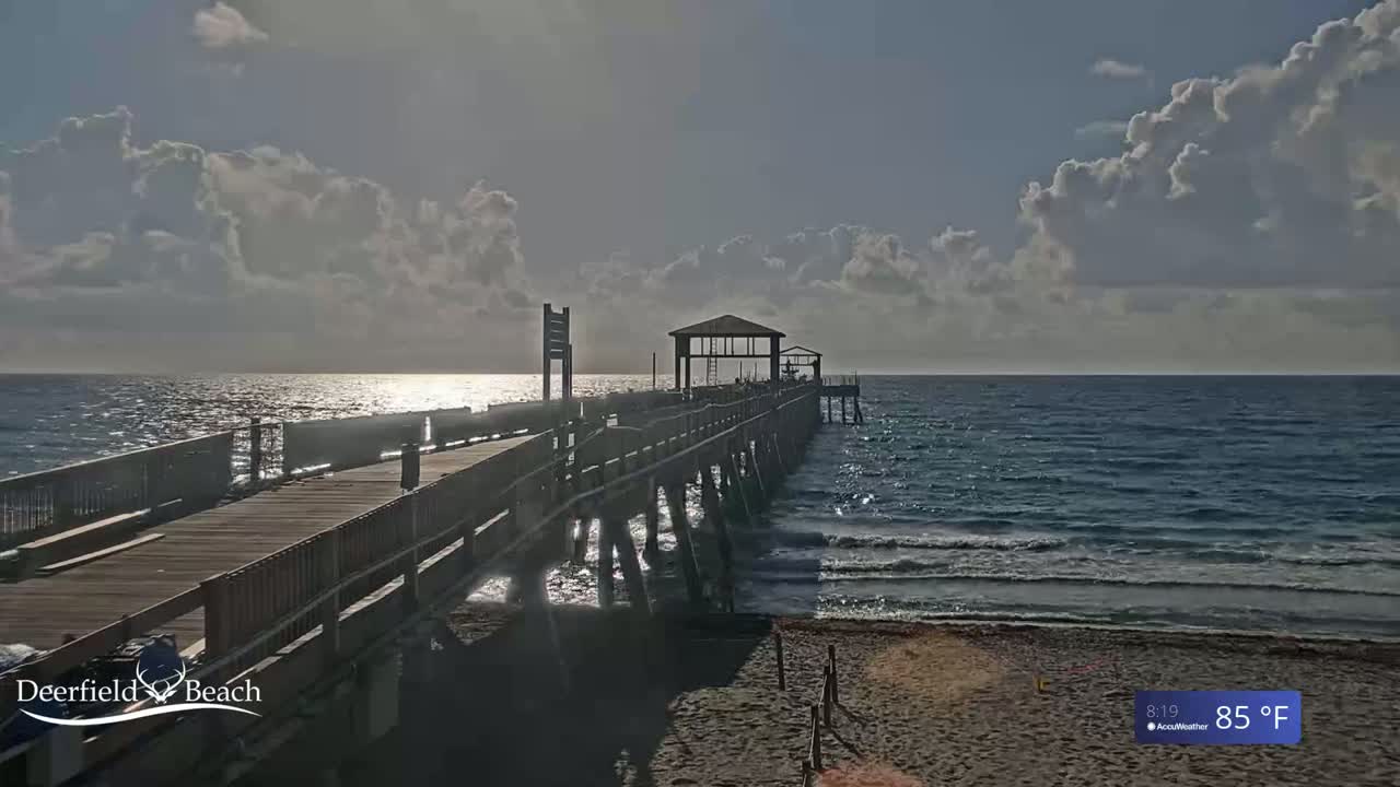 A long wooden pier extends into a calm ocean under a partly cloudy sky, with the sun shining brightly on the water.