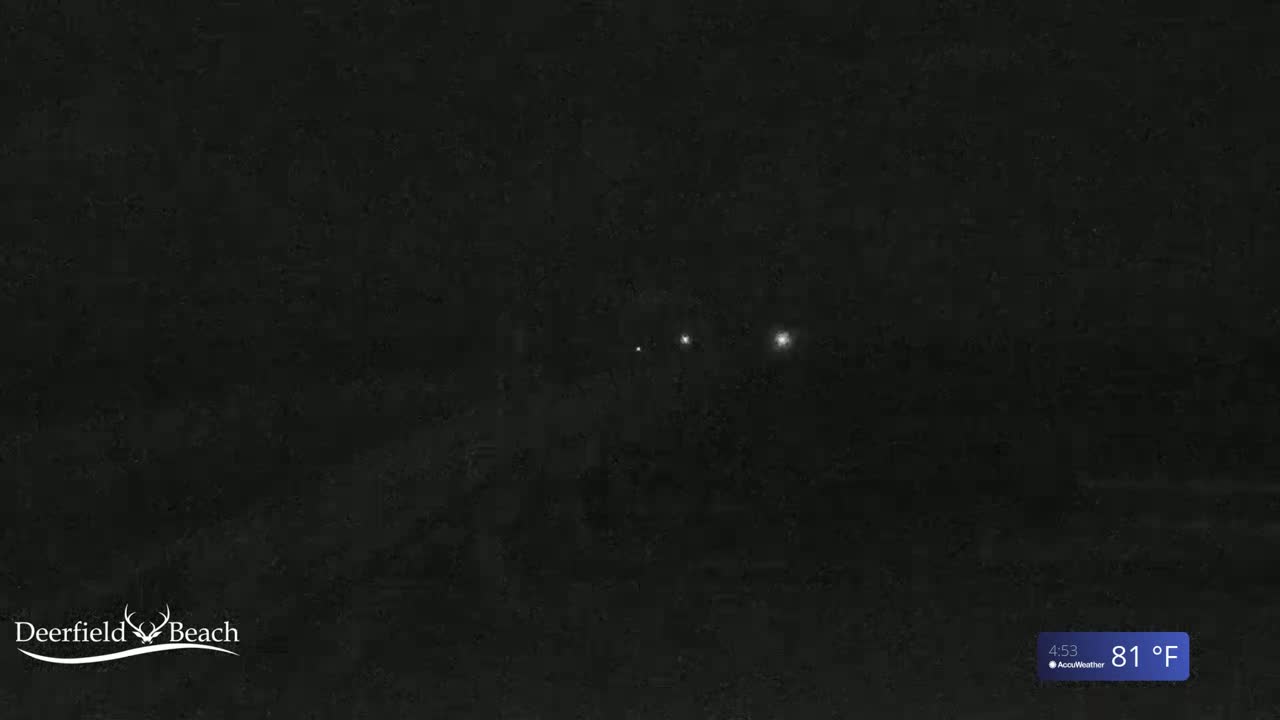 A nighttime scene shows a dark, possibly wooded area with a line of faint lights in the distance under a clear sky.