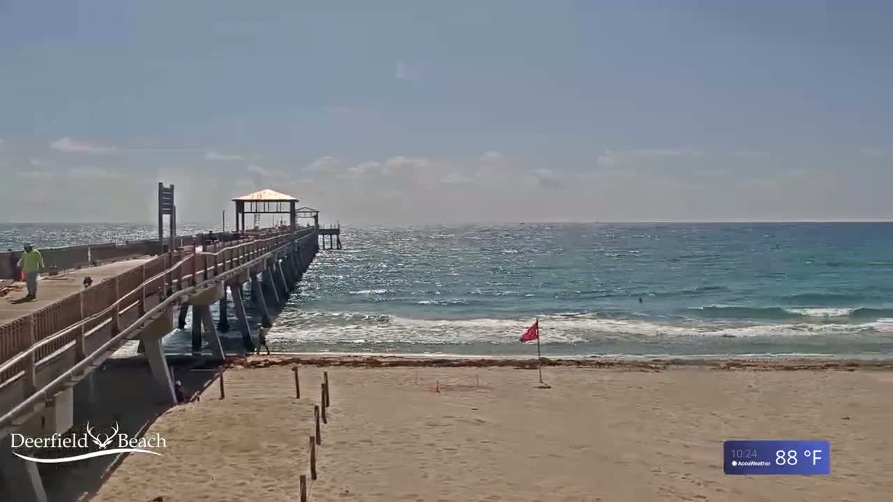 A long pier extends over a sandy beach and turquoise ocean under a mostly sunny sky.