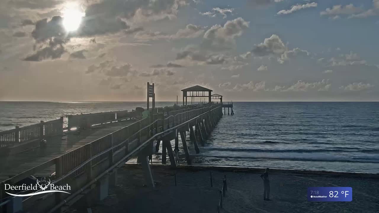 A long pier extends into a calm ocean under a partly cloudy sky, with the sun shining through the clouds.