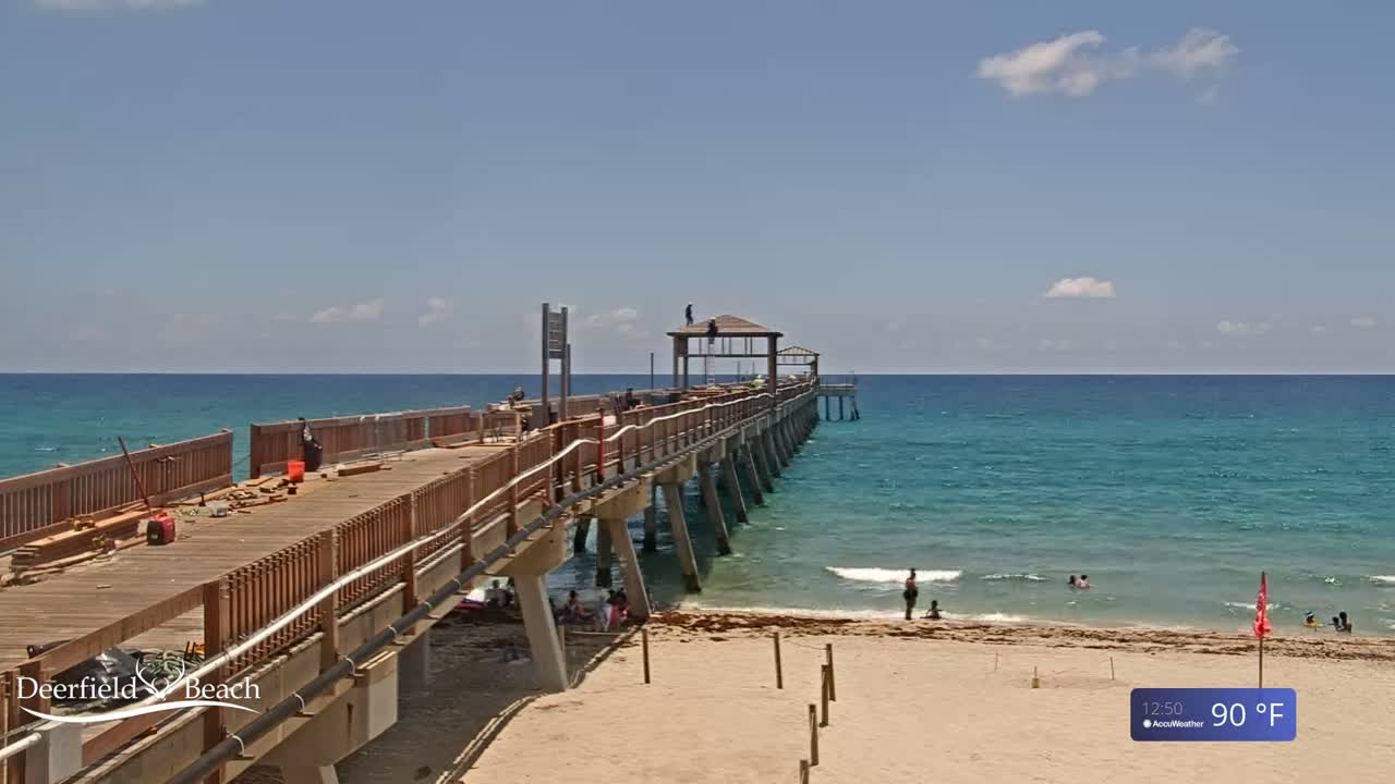 A long wooden pier extends over a calm, turquoise ocean on a sunny day with a few scattered clouds and a temperature of 90°F.