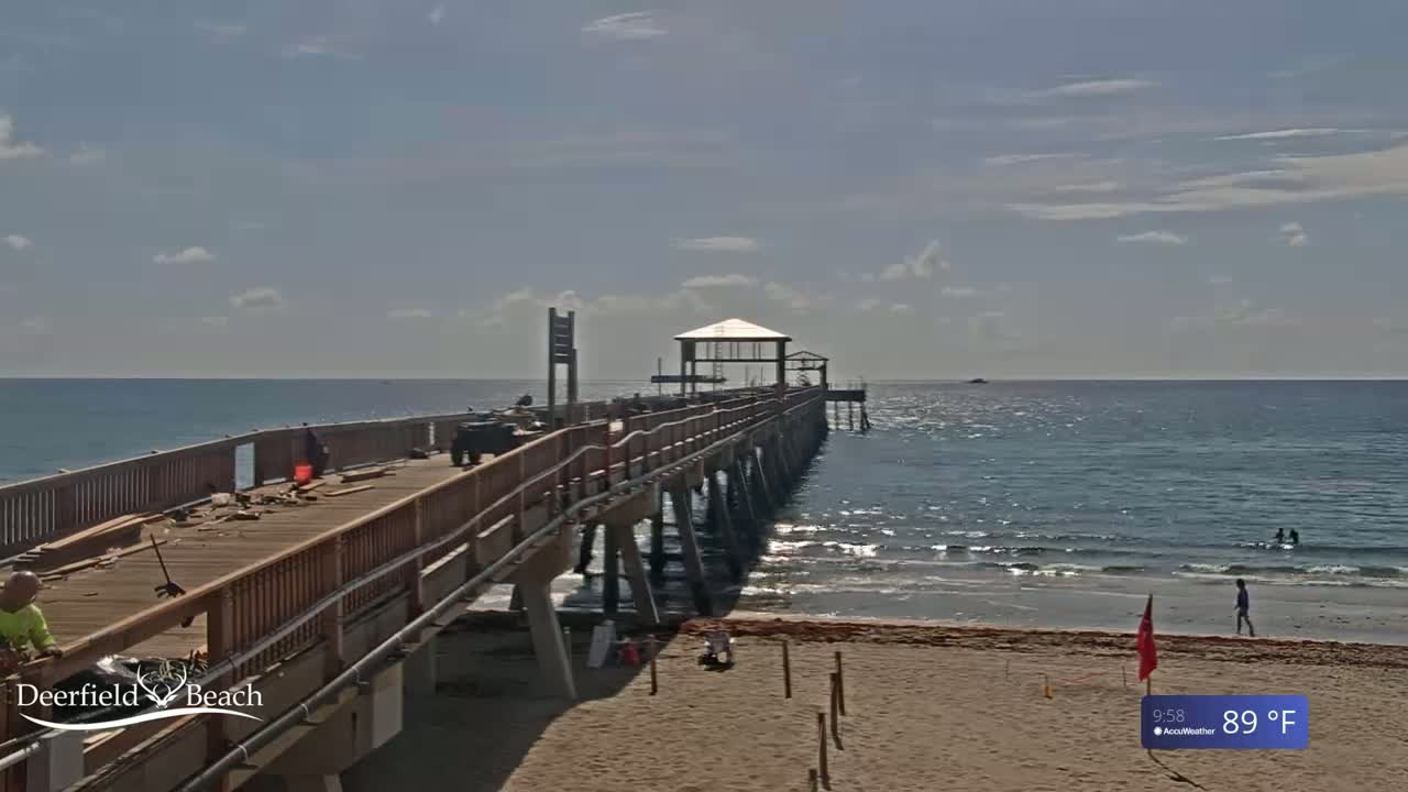 A long pier extends from a sandy beach into a calm ocean under a mostly sunny sky.