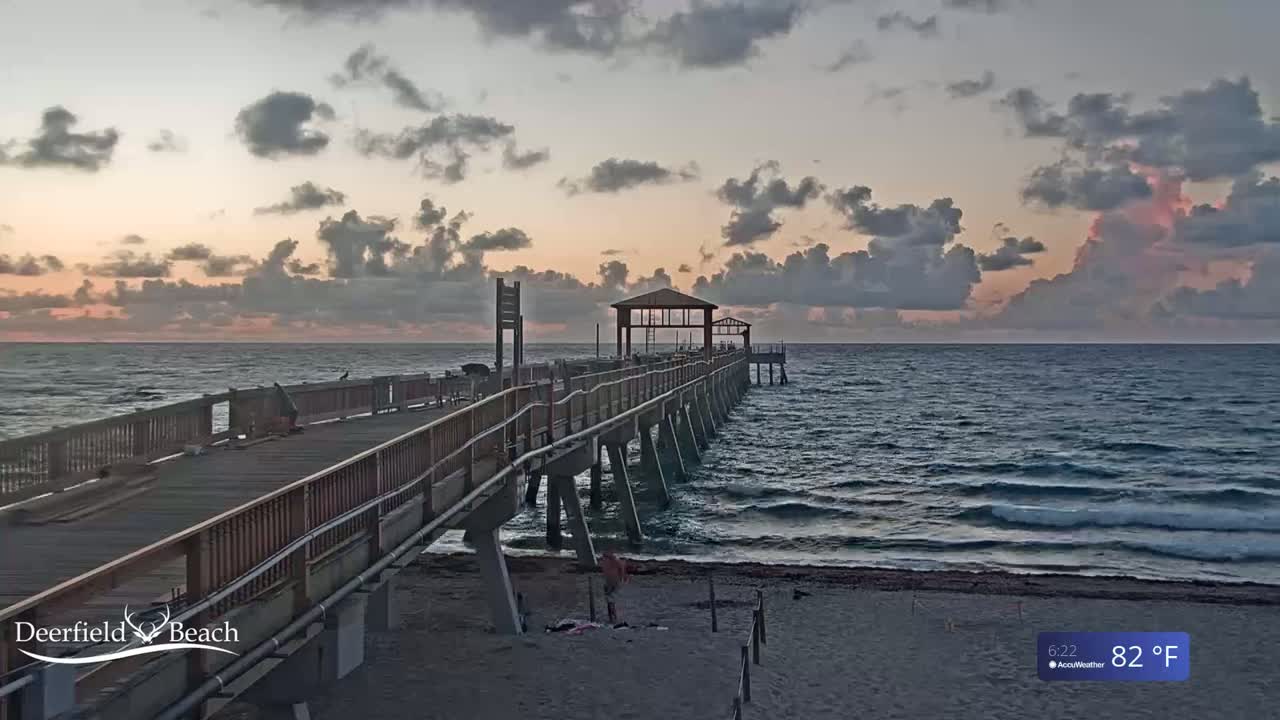 A long pier extends from a sandy beach into a calm ocean under a partly cloudy sky at sunrise.