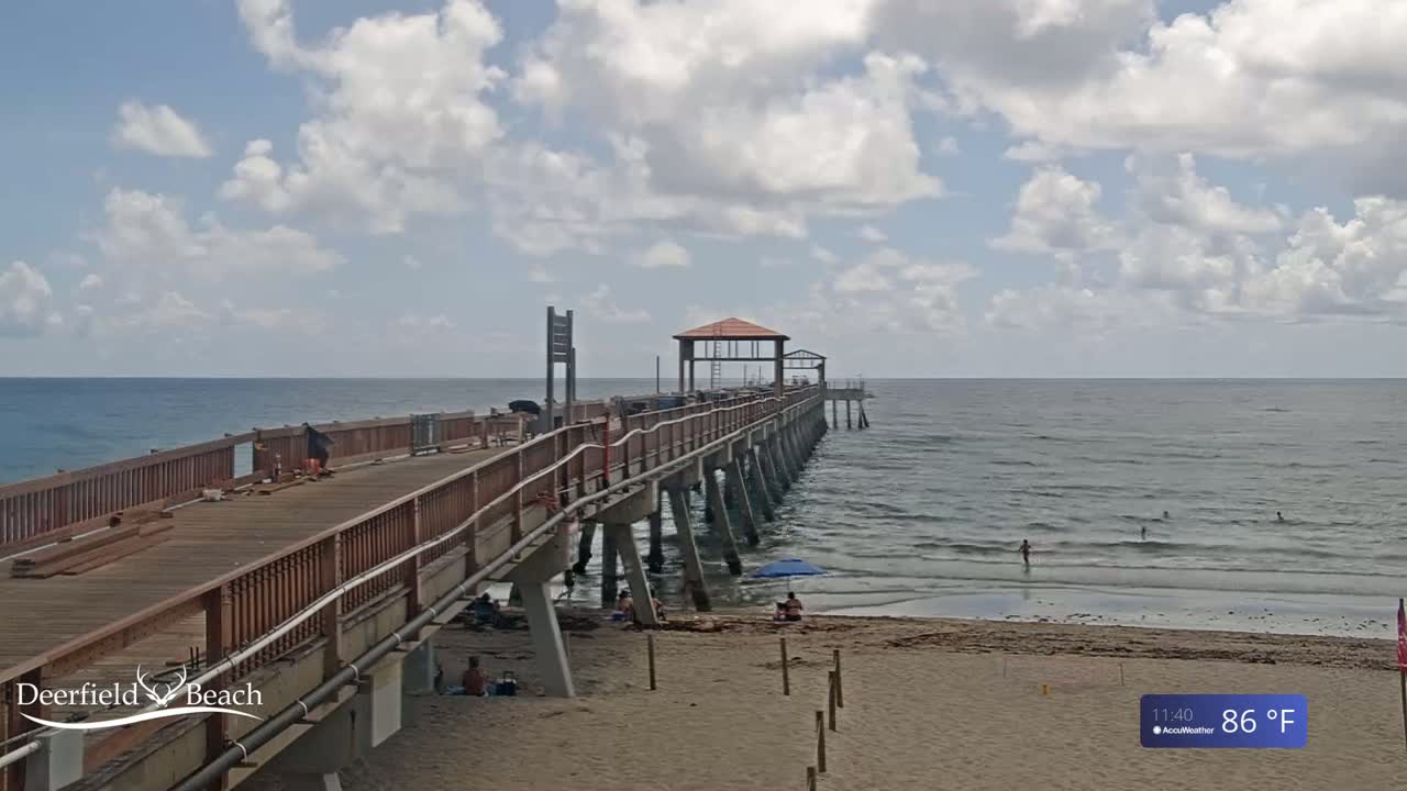 A long wooden pier extends from a sandy beach into a calm ocean under a partly cloudy sky.