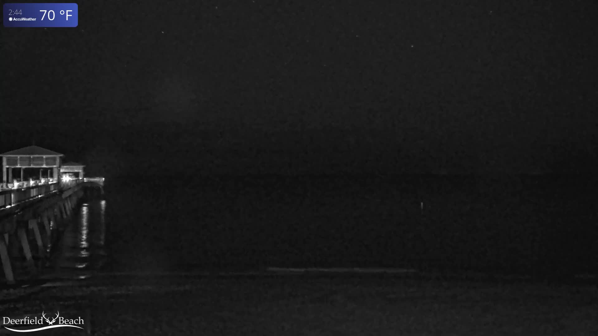 A dark night scene captures a brightly lit pier extending over the ocean under clear skies.