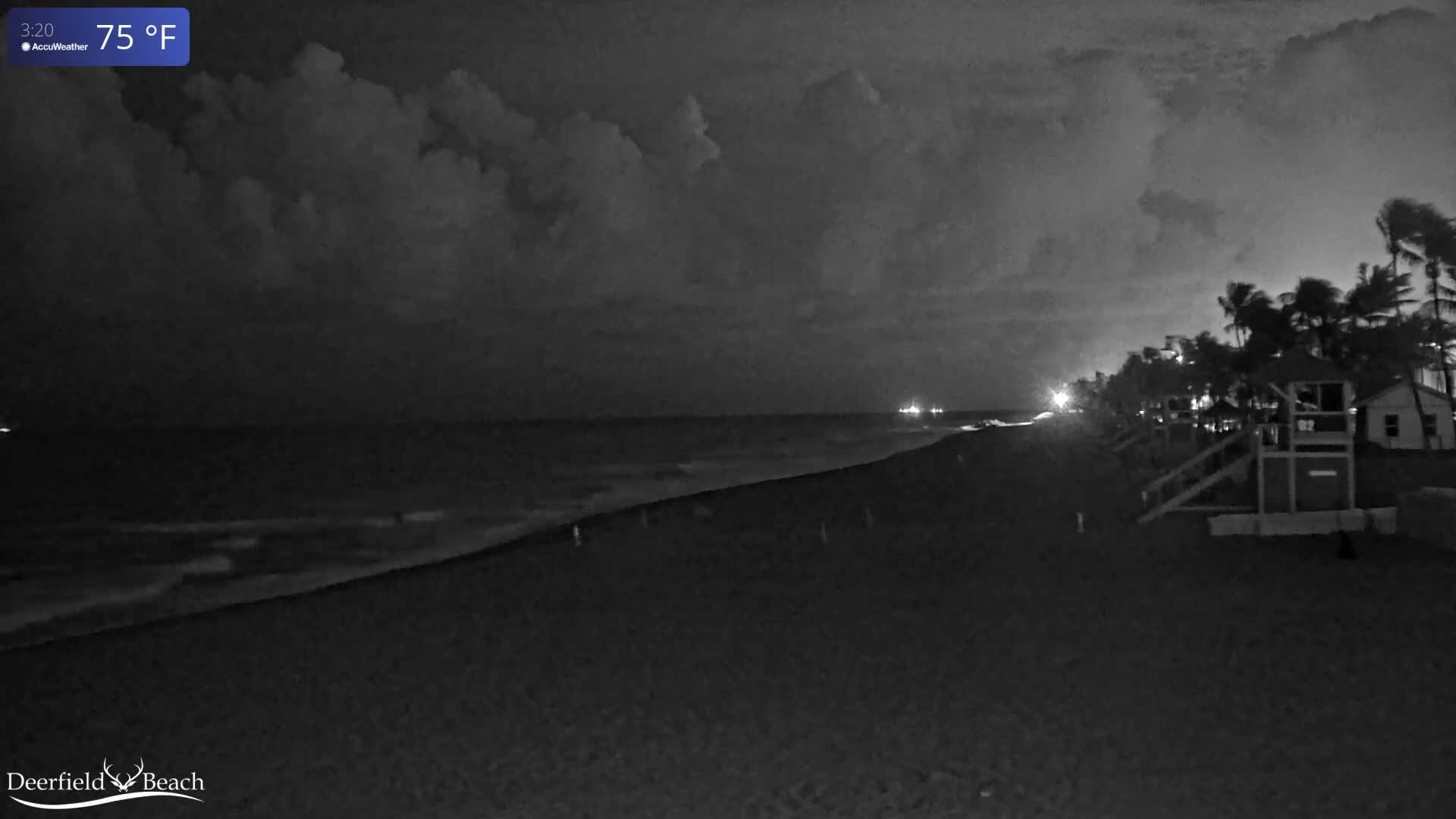 A dark, cloudy night scene shows a beach with gentle waves on the ocean, a sandy shoreline, and distant lights illuminating a line of palm trees and a lifeguard stand.