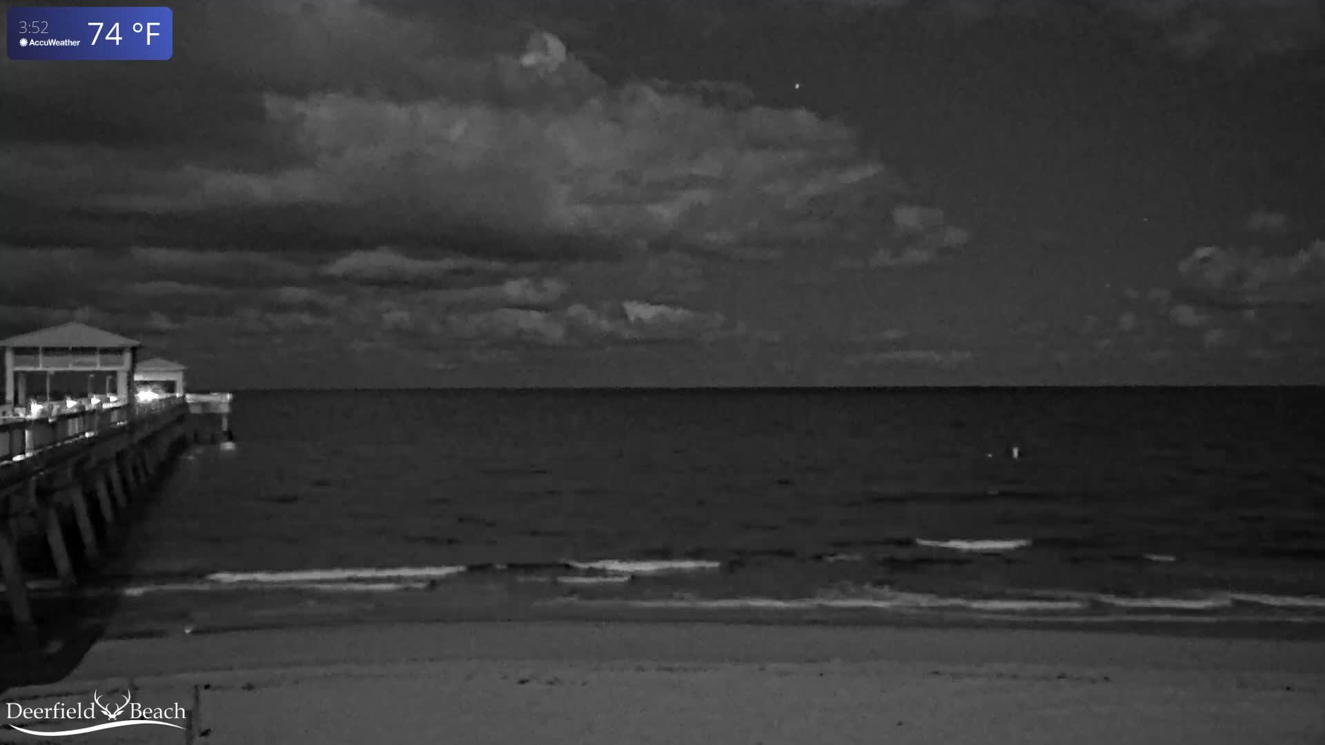 A dark, cloudy night captures a lighted pier stretching into the ocean from a sandy beach, with gentle waves lapping the shore.