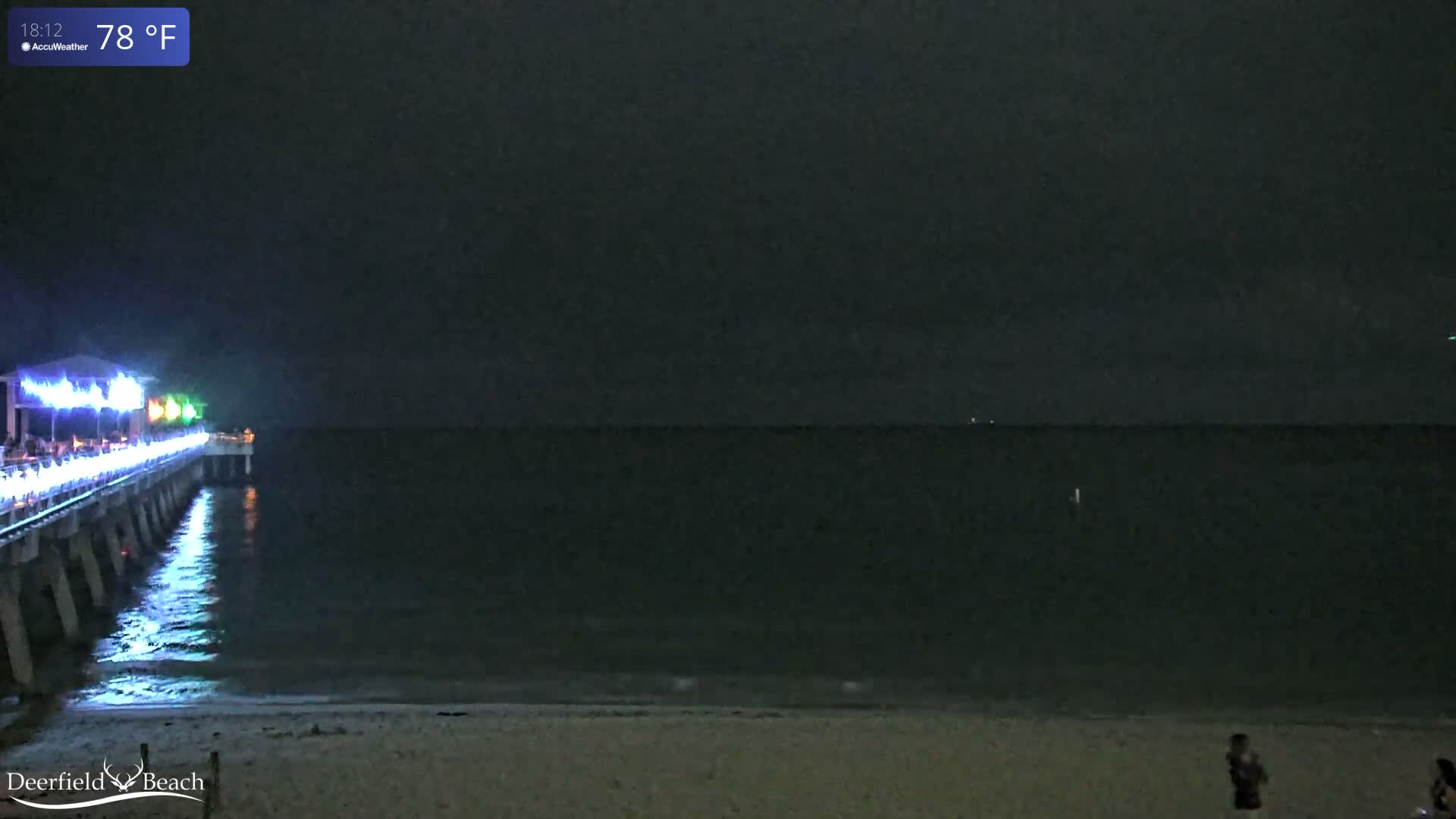 A dark, cloudy night captures a lighted pier stretching into the ocean from a sandy beach, with gentle waves lapping the shore.
