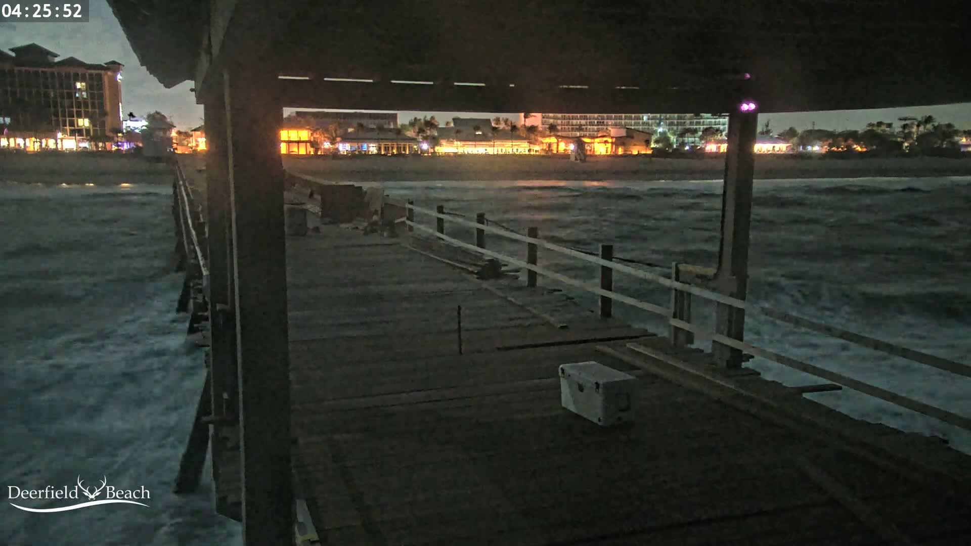 A nighttime view from a pier shows choppy waves crashing against the structure, with illuminated buildings visible in the distance.