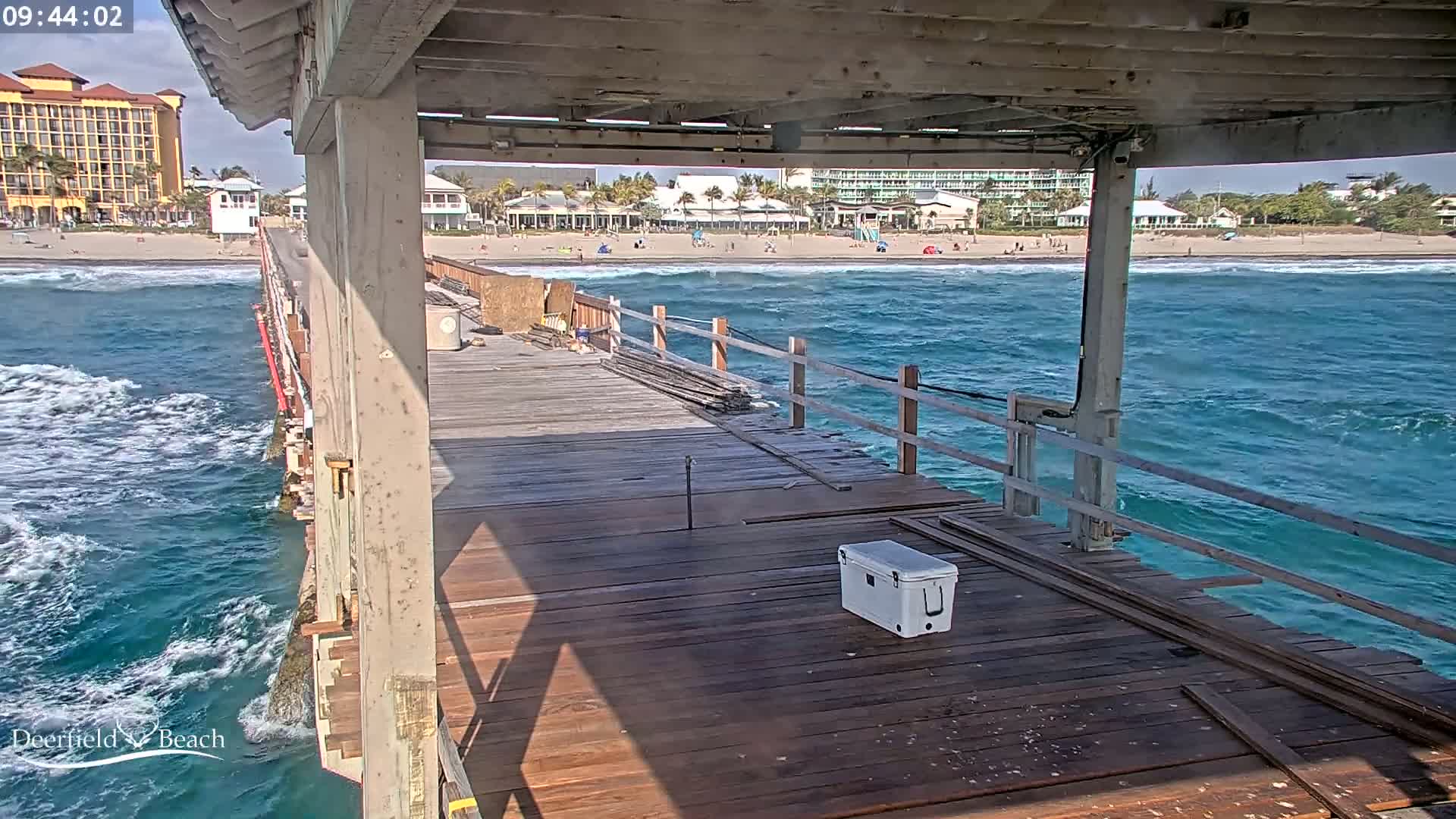 A partly sunny day shows a wooden pier extending over a choppy ocean with a beach and hotels in the background.