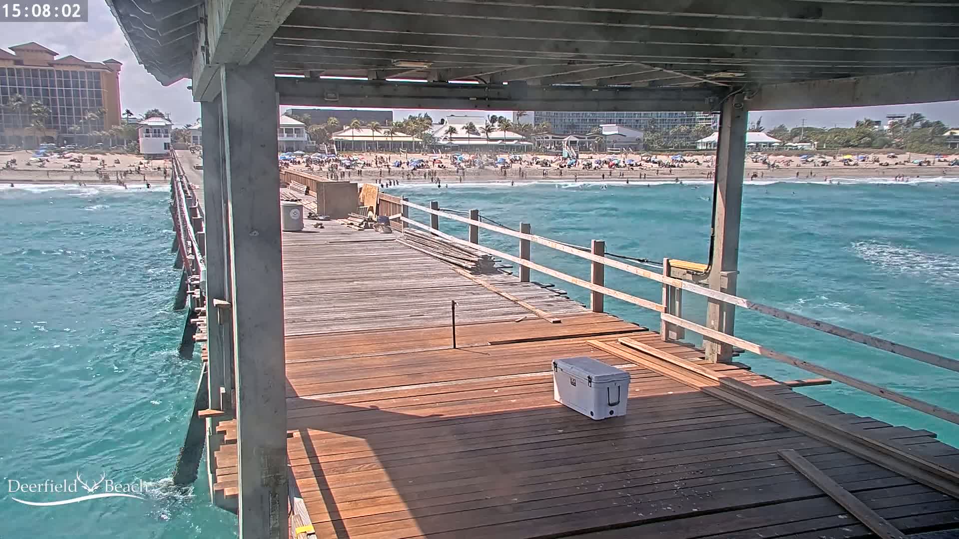 A wooden pier under construction extends over teal ocean water, with a beach and buildings visible in the background on a sunny day.