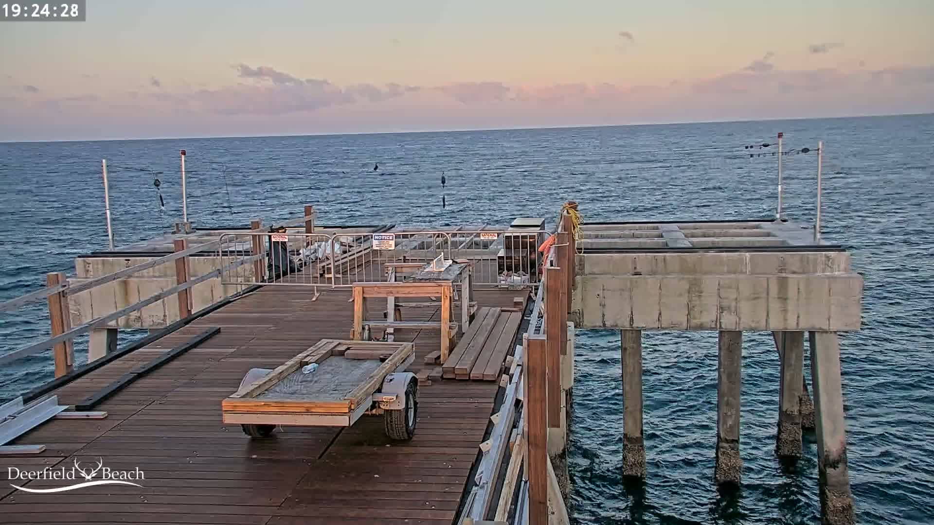 A partially constructed pier extends over a calm ocean under a pale pink and blue twilight sky.