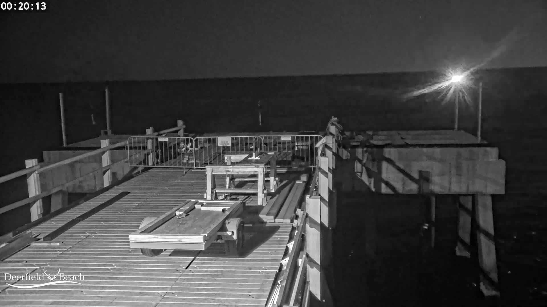 A nighttime, black-and-white view shows a pier under construction with building materials and tools on the deck, overlooking a calm body of water.
